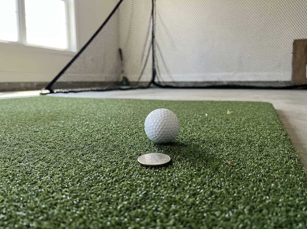 Indoor golf simulator setup featuring a golf ball and coin on artificial turf, with a net in the background, ideal for practicing golf swings and improving skills at home.