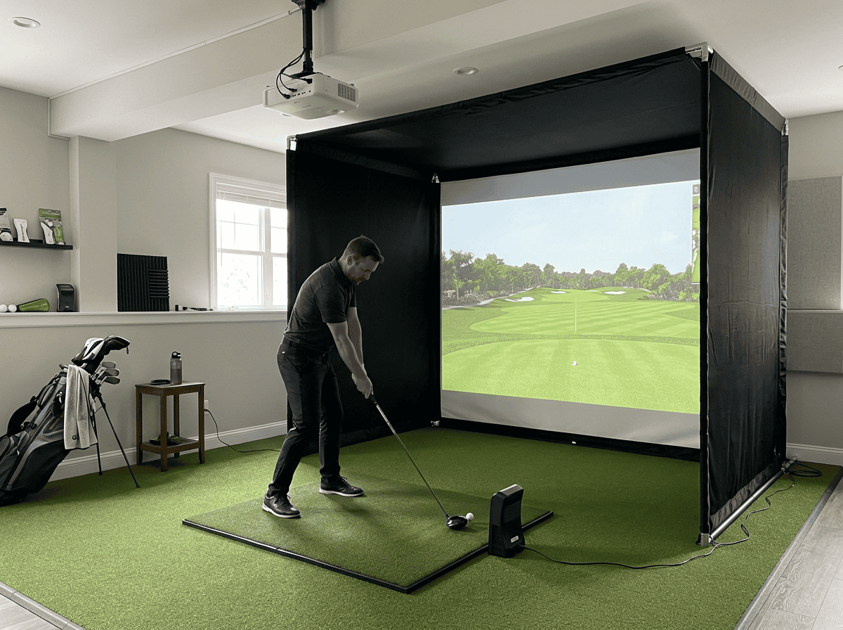 Man practicing golf swing in an indoor golf simulator with a large screen showing a golf course.